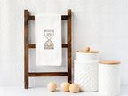A white kitchen towel with a hourglass design embroidered on it. The hourglass is filled with a sun and moon design. The towel is hanging on a wooden ladder shelf with two white ceramic canisters with wooden lids on the bottom shelf.