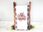 A white kitchen towel with red embroidered text that reads &#39;Feliz Navidad&#39; displayed on a wooden stand with a red and black plaid fabric background.