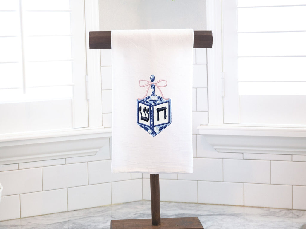 A dreidel in different shades of blue with a pink bow embroidered on a white cotton tea towel set on a towel stand on a white marble kitchen counter with white subway backsplash.
