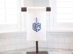 A dreidel in different shades of blue with a pink bow embroidered on a white cotton tea towel set on a towel stand on a white marble kitchen counter with white subway backsplash.