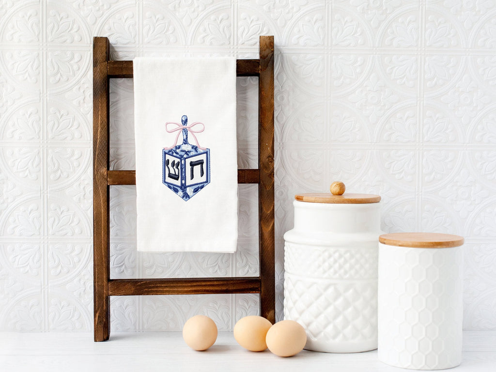 A dreidel in different shades of blue with a pink bow embroidered on a white cotton tea towel set on a towel stand on a white marble kitchen counter with white subway backsplash. There are two white cookie jars and three eggs next to the ladder.