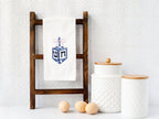 A dreidel in different shades of blue with a pink bow embroidered on a white cotton tea towel set on a towel stand on a white marble kitchen counter with white subway backsplash. There are two white cookie jars and three eggs next to the ladder.