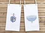 A photo of two white tea towel one with a dreidel in different shades of blue with a pink bow embroidered on a white cotton tea towel and one with a menorah in different shades of blue on a white cotton tea towel. This is showing both towels together with no personalization. The listing is for one towel - with the dreidel with no personalization, but the customer can choose both in the variations section.