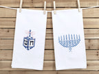 A photo of two white tea towel one with a dreidel in different shades of blue with a pink bow embroidered on a white cotton tea towel and one with a menorah in different shades of blue on a white cotton tea towel. This is showing both towels together with no personalization. The listing is for one towel - with the dreidel with no personalization, but the customer can choose both in the variations section.
