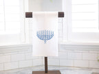 A menorah in different shades of blue with a pink bow embroidered on a white cotton tea towel set on a towel stand on a white marble kitchen counter with white subway backsplash. The menorah embroidered towel is another option in variations that you can choose.