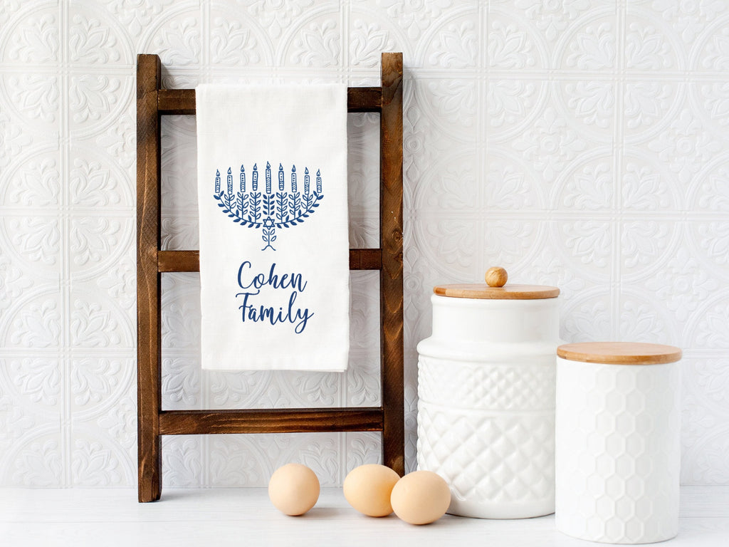 A photo of a dreidel embroidered in different shades of blue with a pink bow embroidered on a white cotton tea towel and set on a ladder. This version of the towel has personalized below: Cohen Family and is an option in variations.