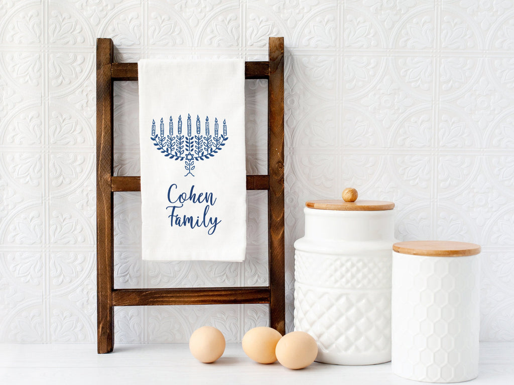 A photo of a dreidel embroidered in different shades of blue with a pink bow embroidered on a white cotton tea towel and set on a ladder. This version of the towel has personalized below: Cohen Family and is an option in variations.