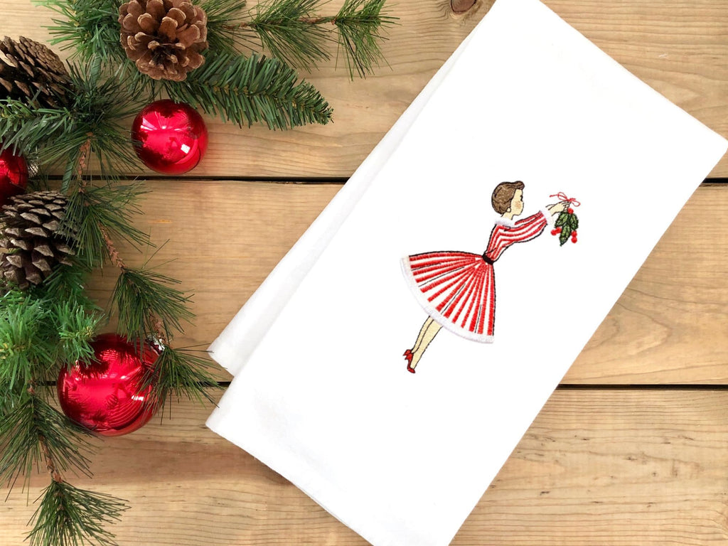 Image of 1950s inspired holiday decor tea towel of a mid century modern woman in a red and white striped dress hanging mistletoe. The tea towel is set on a wooden table with holiday greens and red Christmas ornaments.