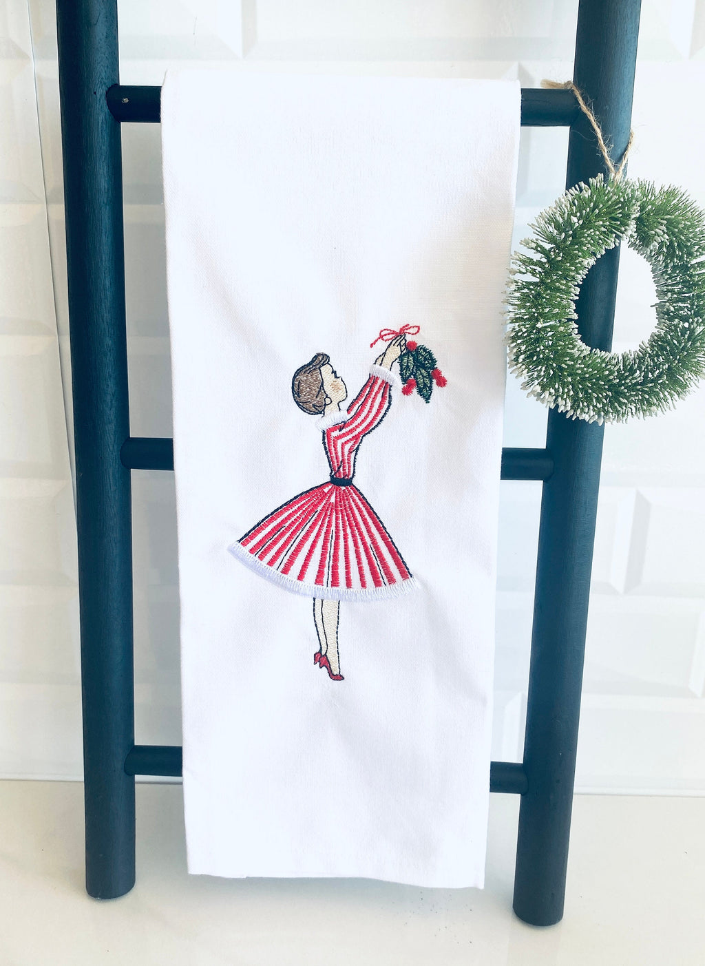 Image of the Meet me under the mistletoe tea towel of a mid century modern woman in a red and white striped dress hanging mistletoe. The tea towel is set on a black towel ladder with a simple wreath on a white counter with white tile backsplash.
