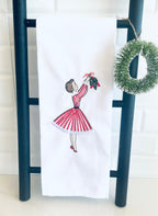 Image of the Meet me under the mistletoe tea towel of a mid century modern woman in a red and white striped dress hanging mistletoe. The tea towel is set on a black towel ladder with a simple wreath on a white counter with white tile backsplash.