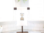 A white kitchen towel with an embroidered hourglass design. The hourglass is filled with gold stars, a sun, and a crescent moon. Set on a white marble countertop with white subway backsplash and windows behind.