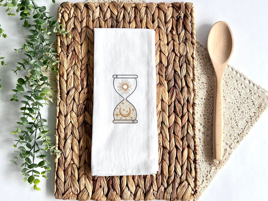 A white kitchen towel with an embroidered hourglass design. The hourglass is filled with a sun and a crescent moon. The towel is on a woven placemat.