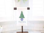 A white kitchen towel with a green Christmas tree in a blue and white pot. The tree is decorated with white lights. The towel has the text &#39;Thomas family&#39; in green script.