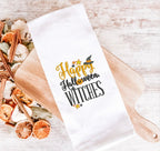 A white tea towel on a light wood cutting board embroidered with the words Happy Halloween Witches with a fall-inspired potpourri at the end of it.