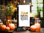 A crisp, white kitchen tea towel embroidered with the words: Happy Halloween Witches in orange and black. The towel in hanging from a tabletop towel display amidst pumpkins and candles.