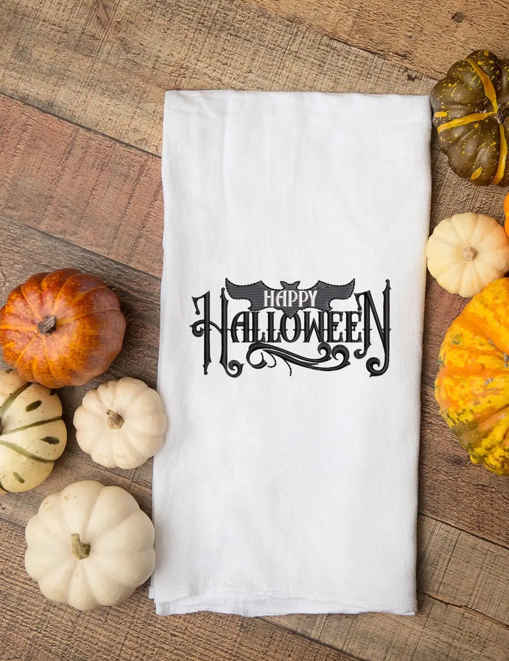 A white kitchen towel with black embroidered text that reads &#39;Happy Halloween&#39; with a bat design in the text. The towel is surrounded by small gourds and pumpkins.