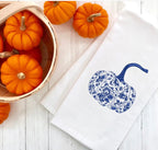 Fall decor chinoiserie blue pumpkin embroidered on a 100%  white cotton tea towel set on a white wooden farmhouse table with a light colored basket filled with mini orange pumpkins.