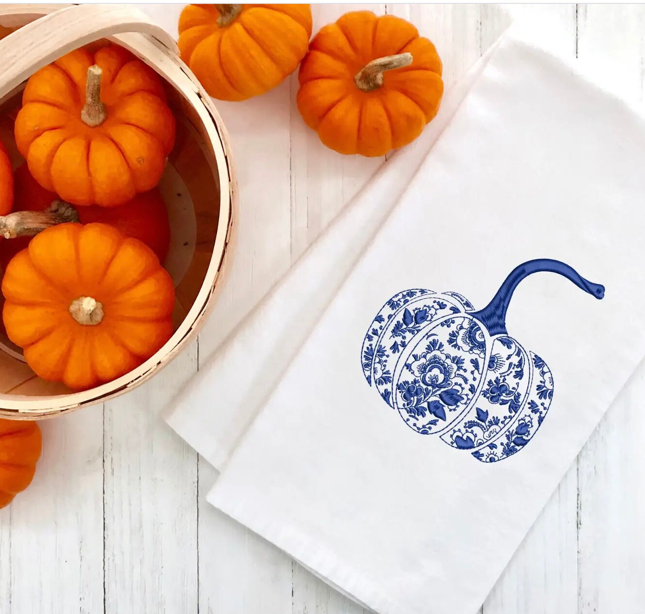 Fall decor chinoiserie blue pumpkin embroidered on a 100%  white cotton tea towel set on a white wooden farmhouse table with a light colored basket filled with mini orange pumpkins.