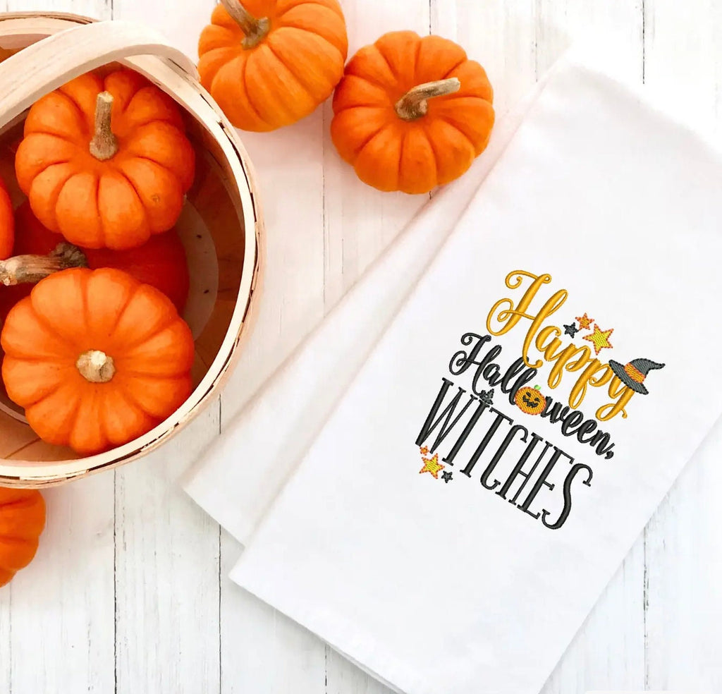 White tea towel embroidered with Happy Halloween Witches in orange and black on a white background next to a basket of small, orange pumpkins.