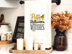 White towel hanging on a kitchen towel rod embroidered with Happy Halloween Witches Set on a wooden tray with candles.