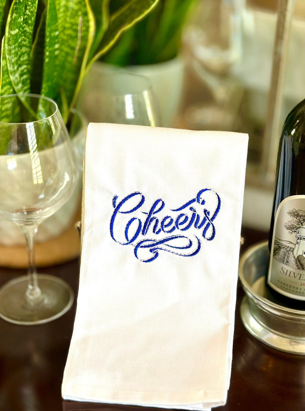 A white towel with a blue &quot;Cheers&quot; embroidered on it. Sitting on a home bar against a couple of wine glasses and next to a bottle of wine.