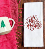 White kitchen towel with red embroidered text that says &#39;Feliz Navidad&#39; set on a brown table with a red fringed table throw and a mug with a Christmas tree next to it.