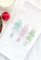 Three embroidered nutcracker figures on a white kitchen towel. The nutcrackers are outlined in green, pink, and blue thread. The nutcrackers are standing in a row and are facing the viewer.