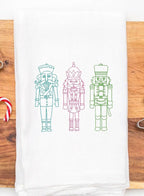 Three embroidered nutcracker figures on a white kitchen towel. The nutcrackers are outlined in green, pink, and blue thread. The nutcrackers are standing in a row from left to right.