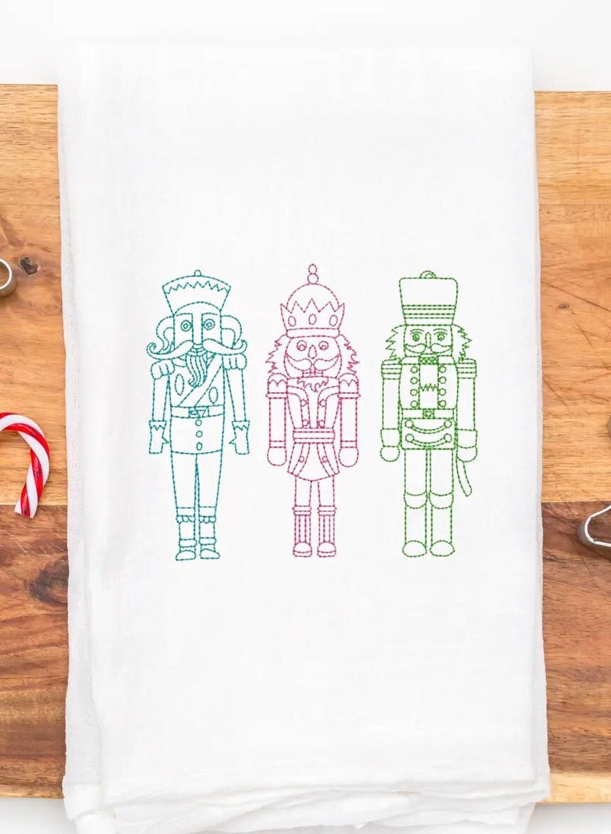 Three embroidered nutcracker figures on a white kitchen towel. The nutcrackers are outlined in green, pink, and blue thread. The nutcrackers are standing in a row from left to right.