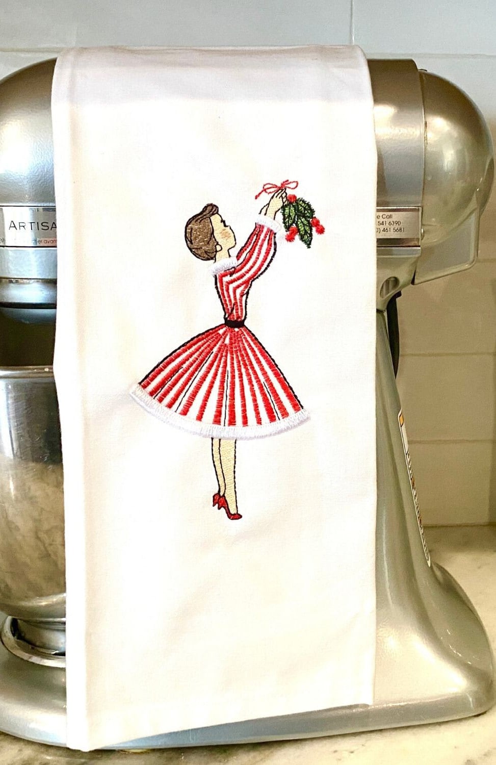 Image of the vintage inspired tea towel of a mid century modern woman in a red and white striped dress hanging mistletoe. The tea towel is set on a kitchen blender on a white counter with white tile backsplash.