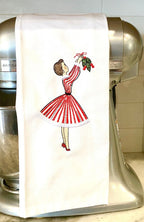 Image of the vintage inspired tea towel of a mid century modern woman in a red and white striped dress hanging mistletoe. The tea towel is set on a kitchen blender on a white counter with white tile backsplash.