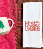 Merry Christmas embroidered in a pretty font where the R&#39;s and S&#39;s have a twirl at the end. Red embroidery. Set on a medium toned wooden table with a red scarf next to it and a white coffee mug with a simple, green painted pine tree on it.