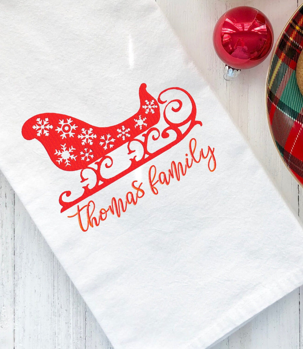 A white kitchen towel with a red sleigh and snowflakes design. The text &#39;thomas family&#39; is embroidered in red below the sleigh.