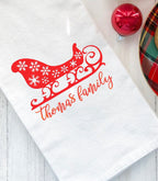 A white kitchen towel with a red sleigh and snowflakes design. The text &#39;thomas family&#39; is embroidered in red below the sleigh.