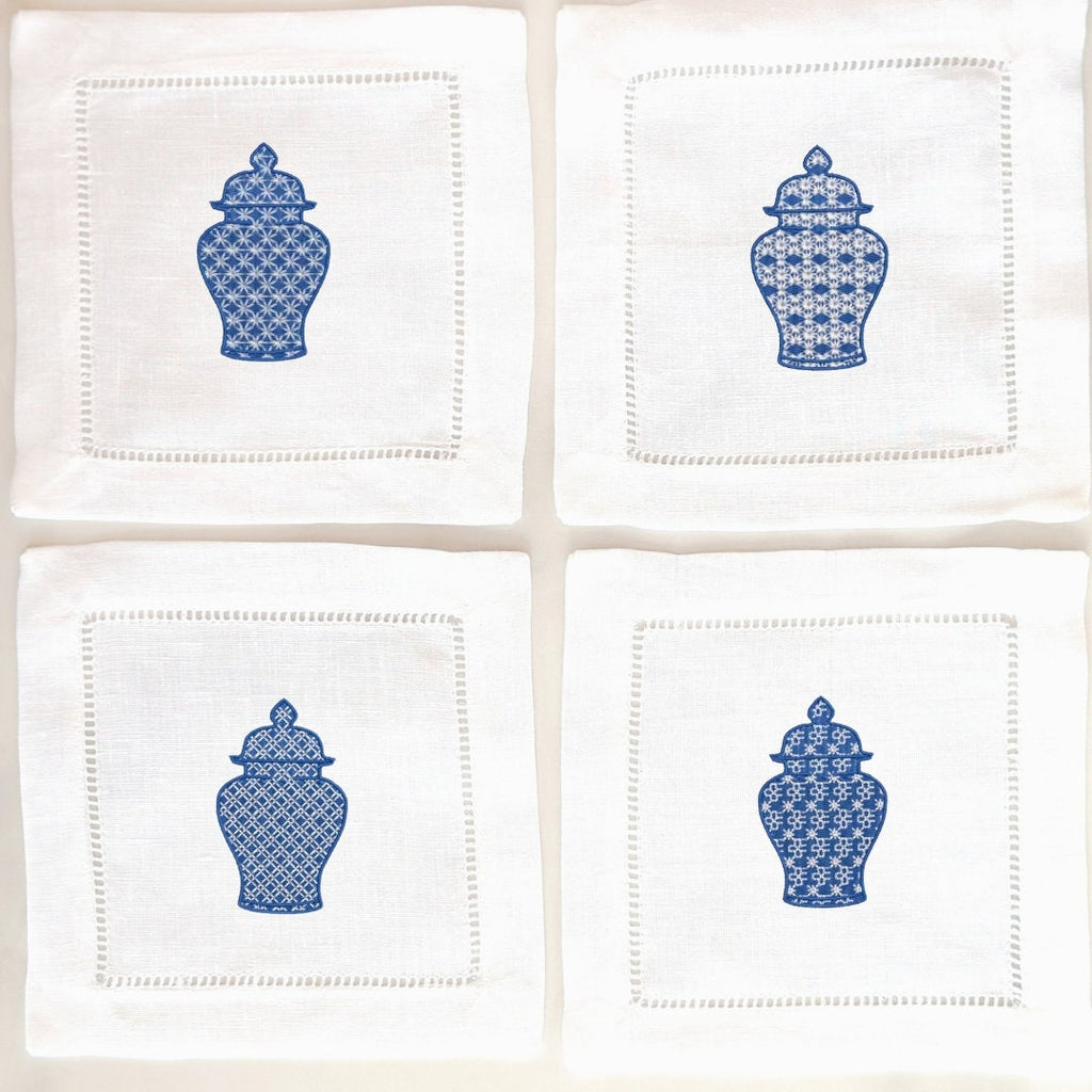 Set of four white linen napkins each with a slightly different chinoiserie design stitched in blue on each. They are set on a white background.