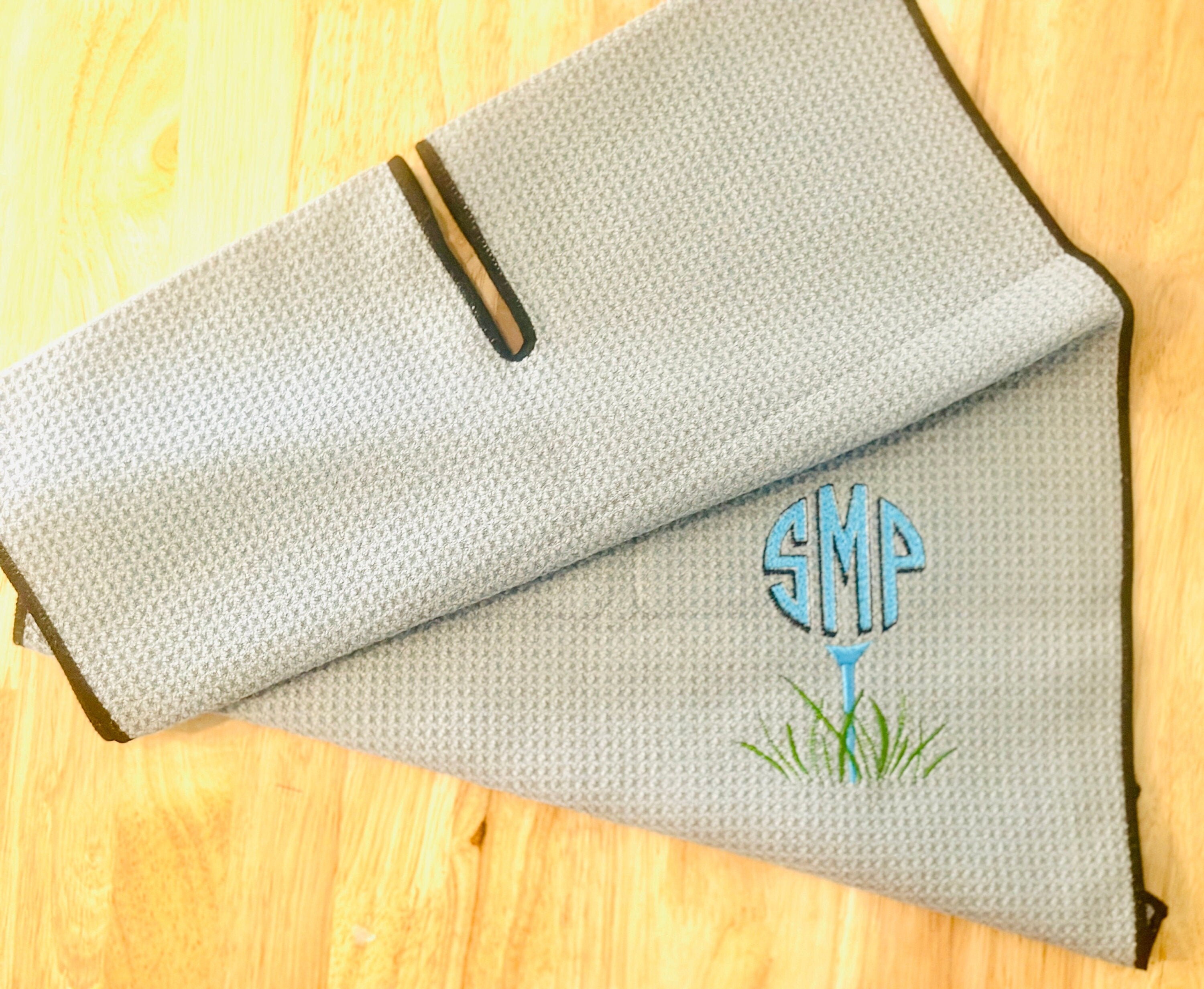 A custom, monogrammed gray waffle weave microfiber golf towel in gray with black trim. The monogram, S M P is in blue and is on top of a tee in the same blue with grass embroidered on the bottom.