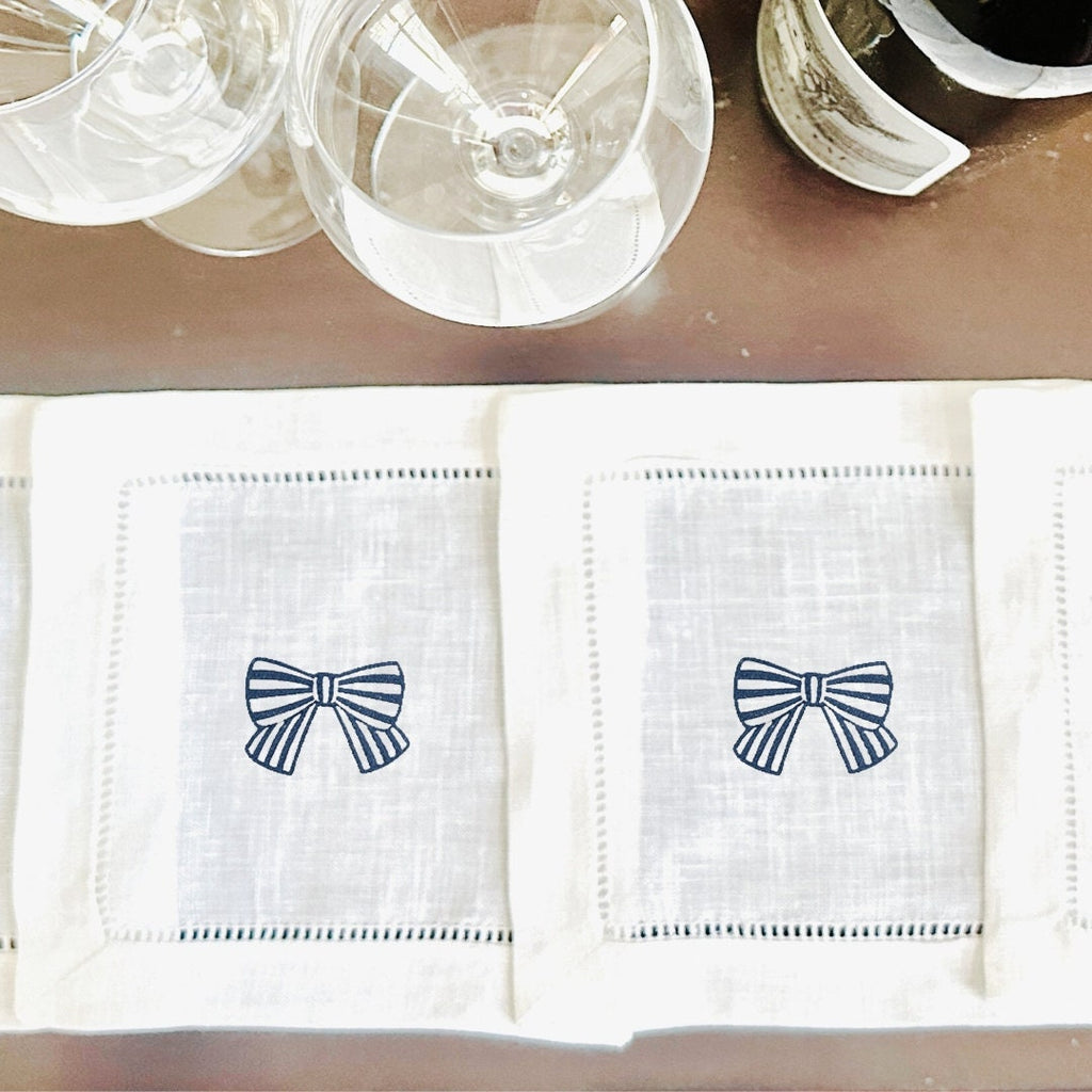 Two white linen napkins with embroidered blue and white bows set on a bar next to wine glasses.