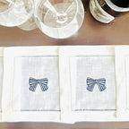 Two white linen napkins with embroidered blue and white bows set on a bar next to wine glasses.