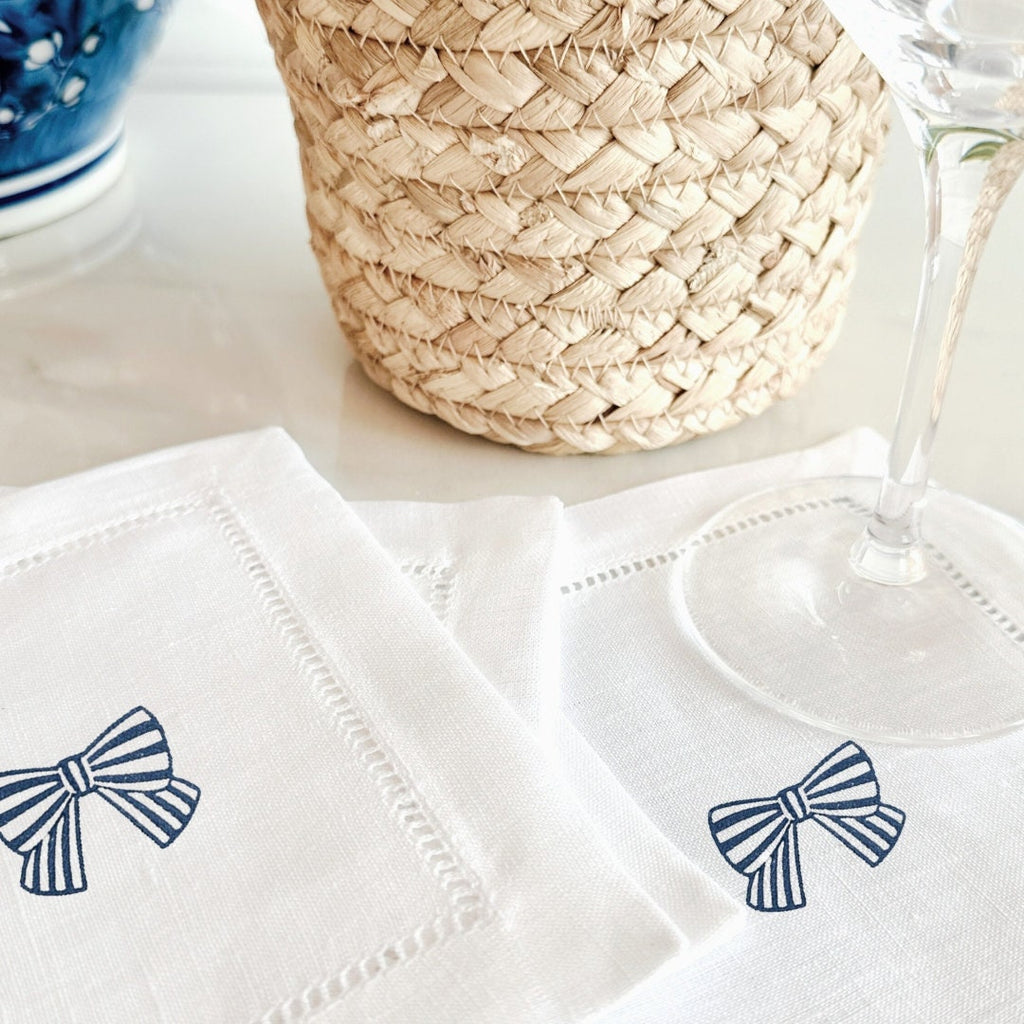 White linen napkins with an embroidered blue and white striped bow set on a white countertop with a basket and a vase nearby. There is a single wine glass set upon a napkin.