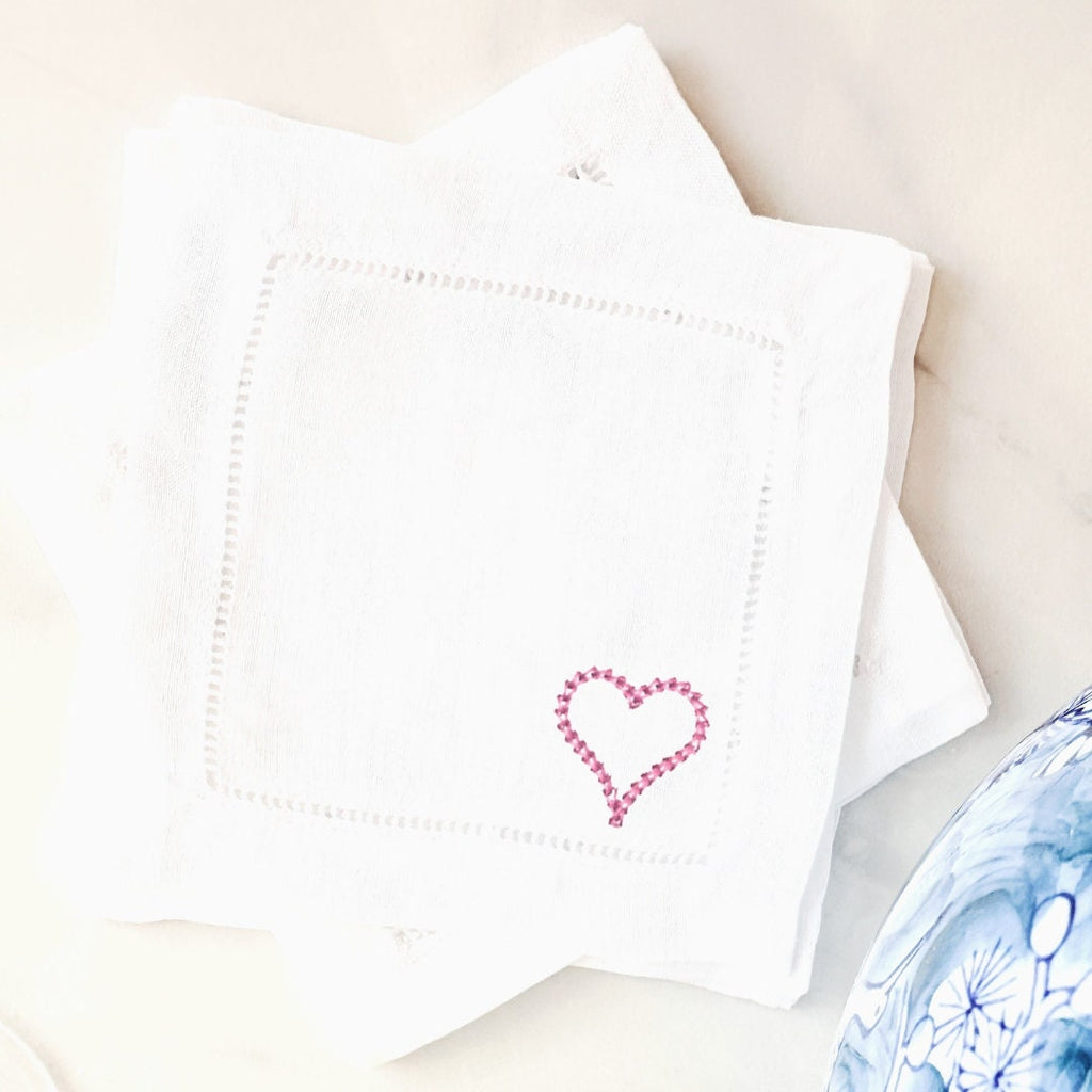 A pink heart embroidered on the bottom right of a white linen cocktail napkin perfect for wedding rehearsal dinner favor set on a white marble counter with a chinoiserie vase to the bottom right.