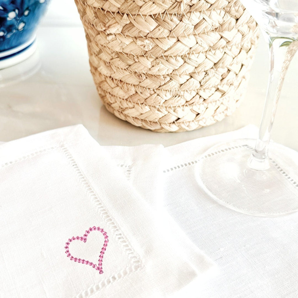 Embroidered pink heart on a white linen cocktail napkin on a marble countertop with a wineglass and wicker basket.