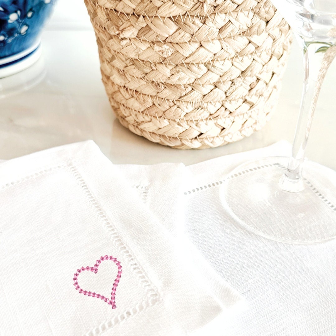 Embroidered pink heart on a white linen cocktail napkin on a marble countertop with a wineglass and wicker basket.