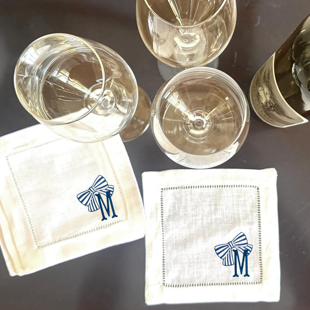 Two white linen cocktail napkins with blue and white striped bows and the letter &#39;M&#39; embroidered on them. The napkins are on a dark brown surface with two clear wine glasses and a bottle of wine.