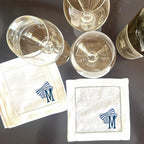 Two white linen cocktail napkins with blue and white striped bows and the letter &#39;M&#39; embroidered on them. The napkins are on a dark brown surface with two clear wine glasses and a bottle of wine.
