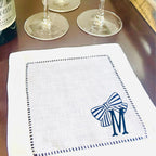 A white linen napkin on a dark table. The napkin has a blue and white striped bow and the letter &#39;M&#39; embroidered in the center of the bow.