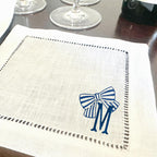 A white linen napkin with a blue and white striped bow and the letter &#39;M&#39; embroidered on it.