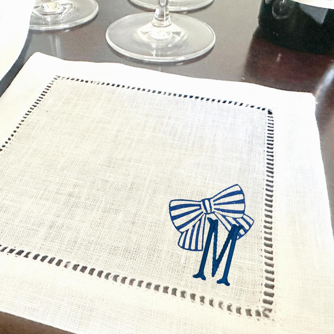 A white linen napkin with a blue and white striped bow and the letter &#39;M&#39; embroidered on it.