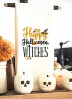 A bright white tea towel with the words Happy Halloween Witches embroidered on it hanging from a black tea towel stand with white pumpkin skulls next to orange mums in a white ceramic vase.