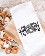 White tea towel with Happy Halloween on a cutting board with an apple, orange, cinnamon potpourri next to it. The stitching saying &quot;Happy Halloween&quot; is done all in black.