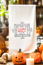 &quot;The Witch is In&quot; embroidered on a white tea towel. Sweet Humble Home HalloweenTea Towel surrounded by jack o lanterns